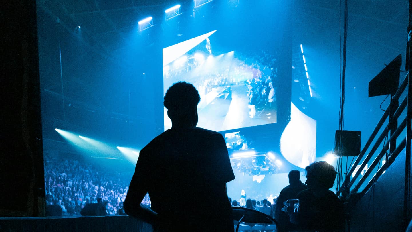 Artista de espaldas mirando al público desde el escenario, con pantallas encendidas